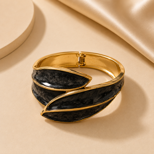 A gold-toned wrap-style cuff bracelet with textured black inlay panels, photographed on a warm beige surface with soft satin fabric in the background.