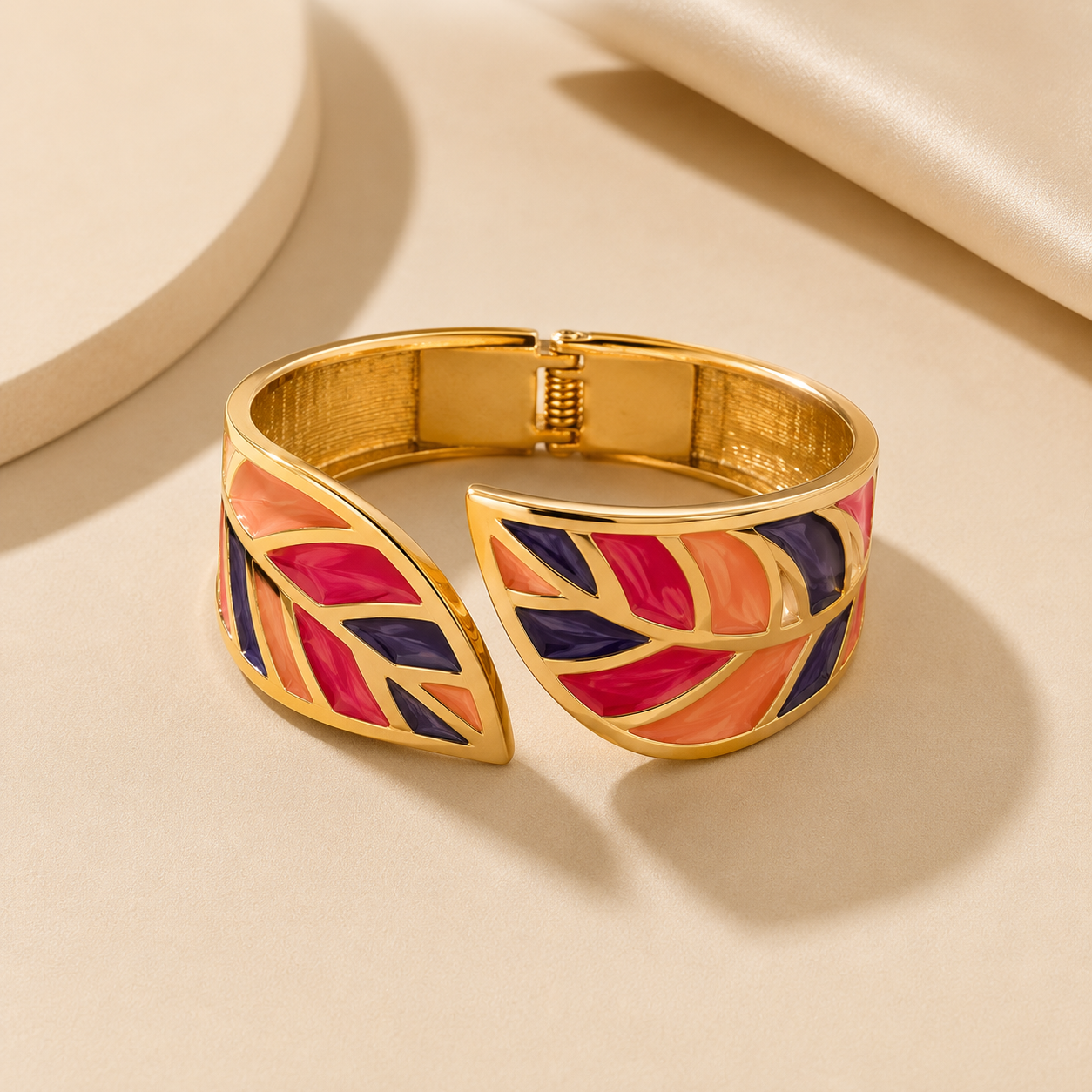 Gold-toned cuff bracelet with a leaf motif in vibrant pink, orange, and navy blue enamel, displayed on a warm beige surface.