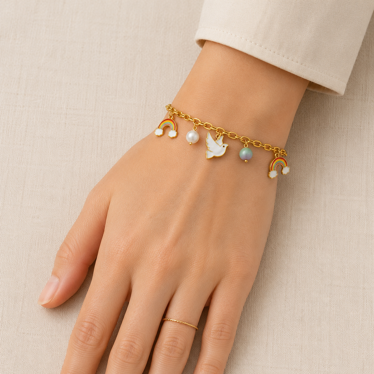 Gold plated rainbow and dove charm bracelet with pearl accents worn on a woman's wrist against a beige background.