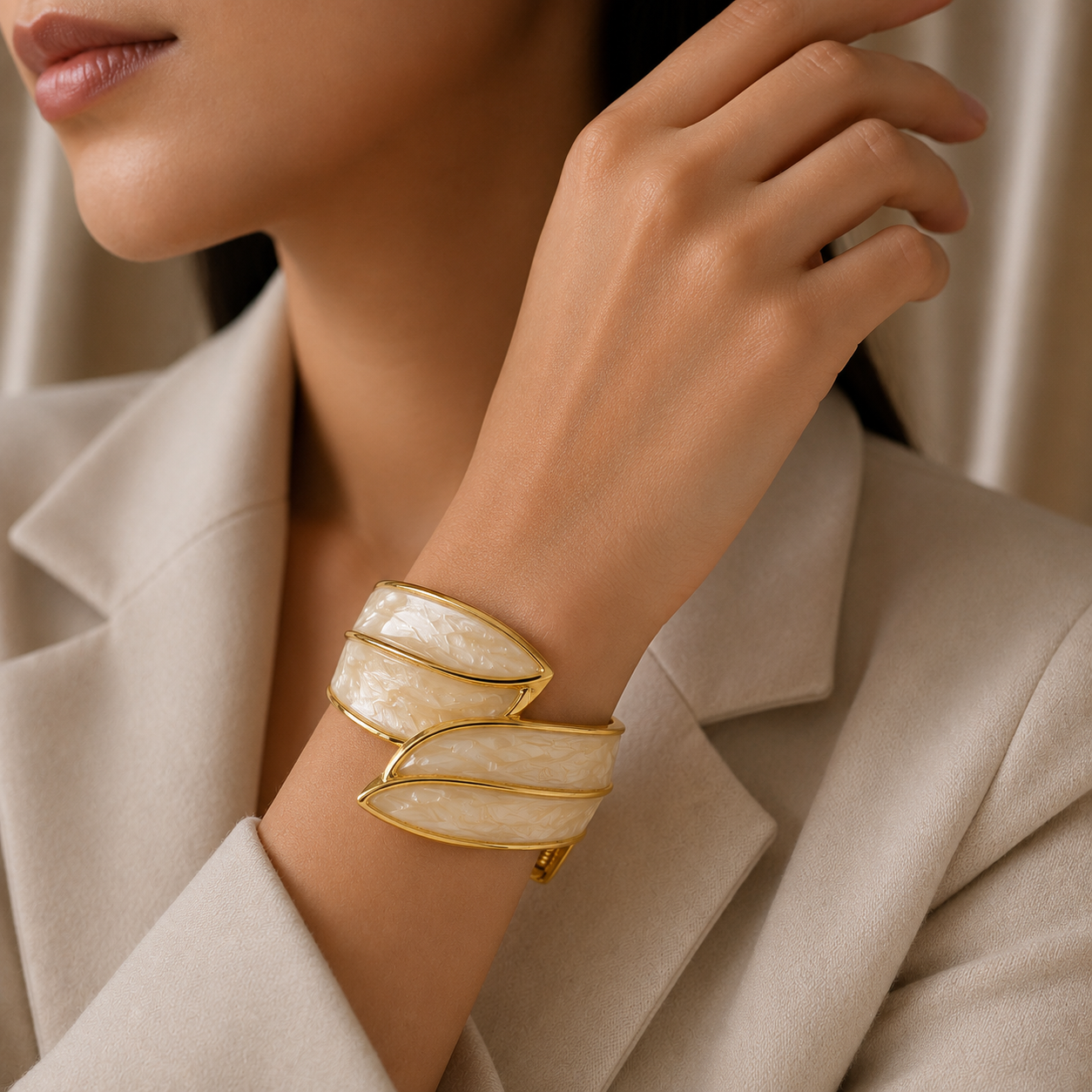 A close-up of a woman wearing a wide gold cuff bracelet with layered, curved off-white mother-of-pearl inlays, styled with a beige blazer.