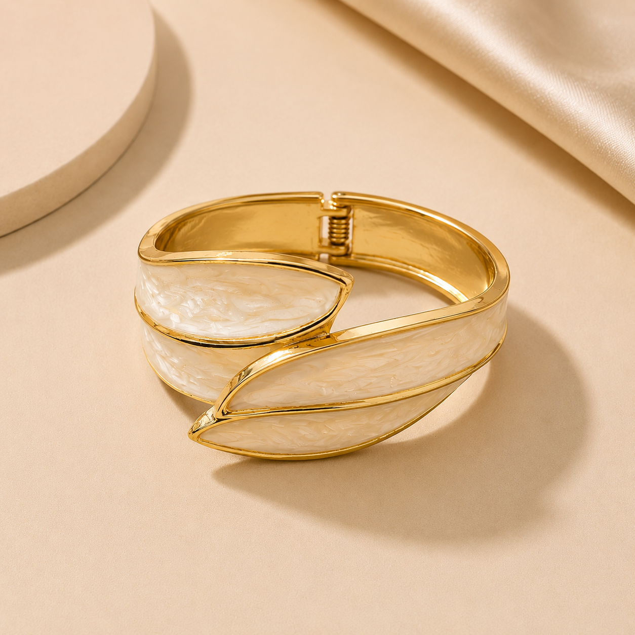 A gold-toned wrap cuff bracelet with smooth, curved white mother-of-pearl inlay panels, styled on a warm beige surface with soft satin fabric in the background.