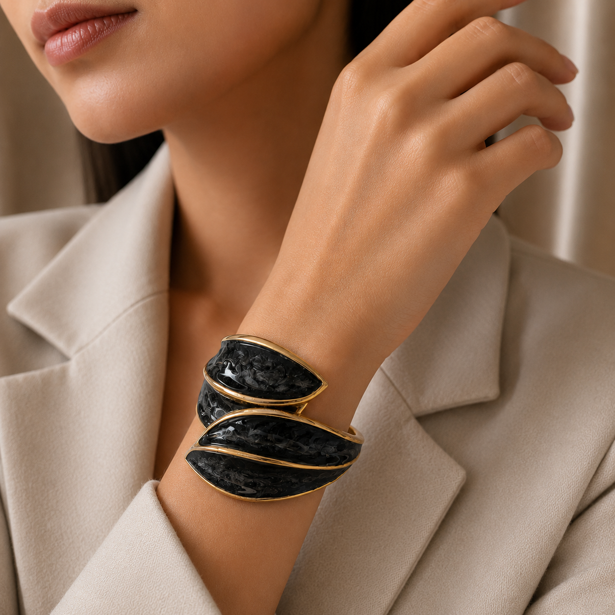 A close-up of a woman wearing a wide wrap-style cuff bracelet with black textured inlay and gold-tone edges, paired with a beige blazer.
