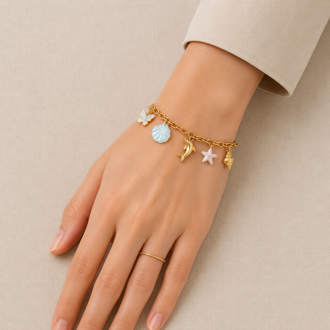 Ocean-inspired gold-tone charm bracelet featuring butterfly, blue seashell, dolphin, pink starfish and spiral shell charms displayed on a premium cream linen background.