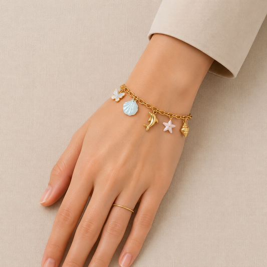 Ocean-inspired gold-tone charm bracelet featuring butterfly, blue seashell, dolphin, pink starfish and spiral shell charms displayed on a premium cream linen background.
