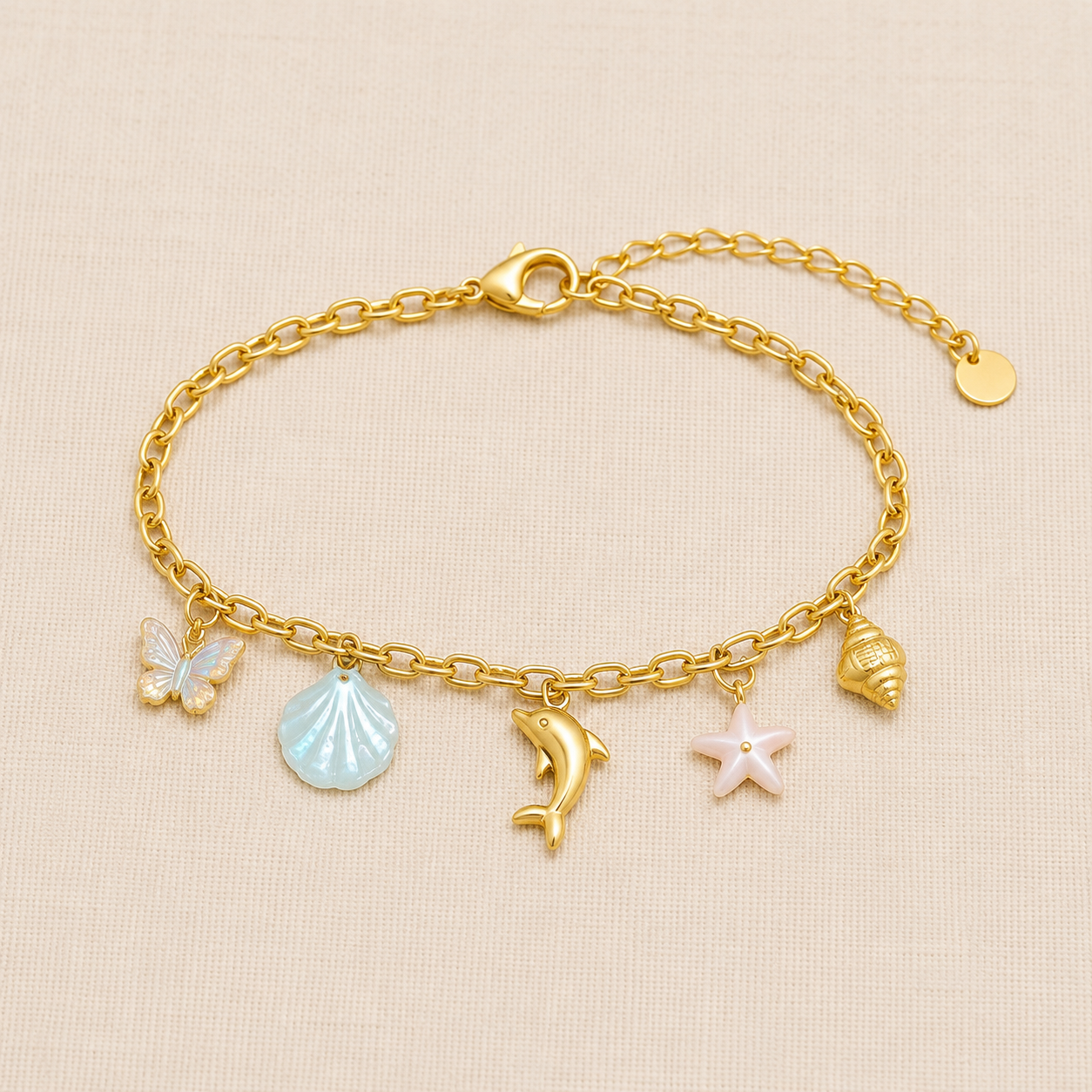 Ocean-inspired gold-tone charm bracelet featuring butterfly, blue seashell, dolphin, pink starfish and spiral shell charms displayed on a premium cream linen background.
